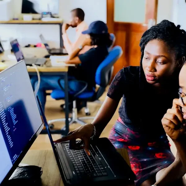 Two engineers debugging code together on a laptop in a busy office, illustrating the complexity organisations face when operating models cannot keep pace with AI